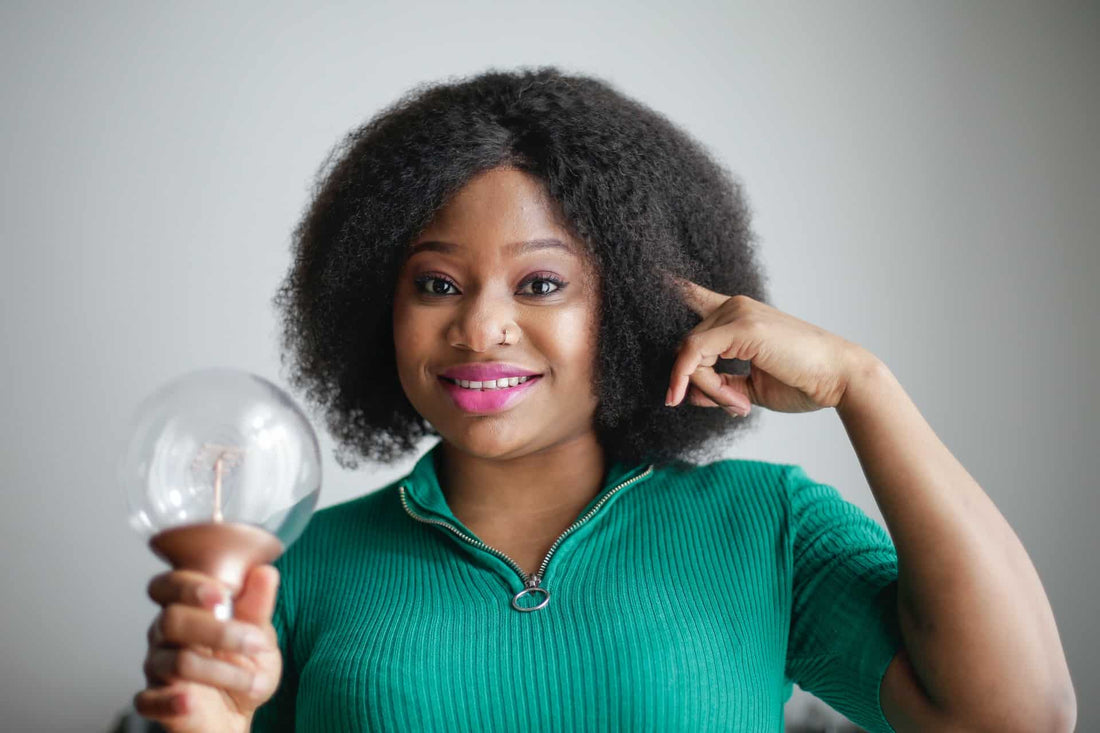 A woman holding a lightbulb and pointing to her head with a smile.